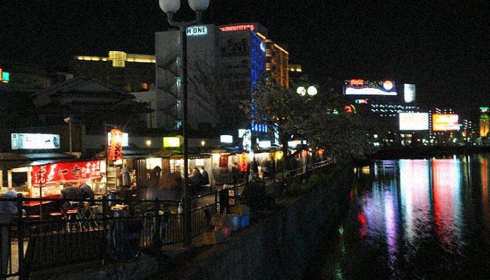Nakasu island in Fukuoka, Japan, with open-air food stalls along the city river and glowing ads on the background that reflect on water, to demonstrate the cyberpunk side